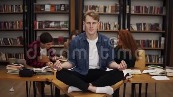 在图书馆学习期间年轻的男学生在桌上打坐他的同学在桌上摆姿势视频的预览图
