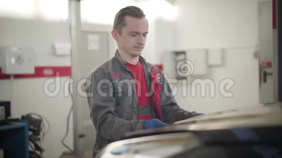年轻自信的白种人的肖像穿着工作服打开汽车引擎盖看着里面专业汽车技师视频的预览图