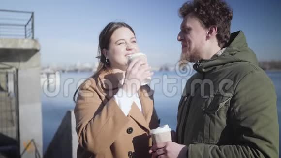 一对快乐的白种人年轻夫妇在河岸上聊天喝咖啡微笑的胖乎乎的肖像视频的预览图