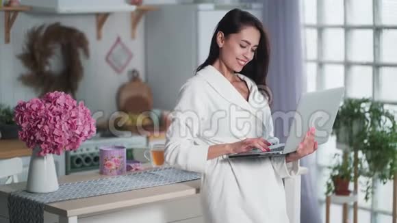 年轻女性穿着白色浴袍的自由职业者在家厨房里用笔记本电脑工作视频的预览图