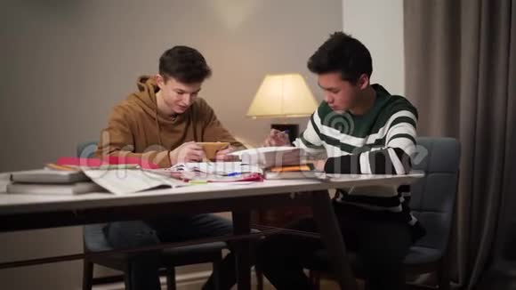两个大学生坐在桌子旁拿着书和工作簿作为白人朋友学习的亚裔男孩视频的预览图