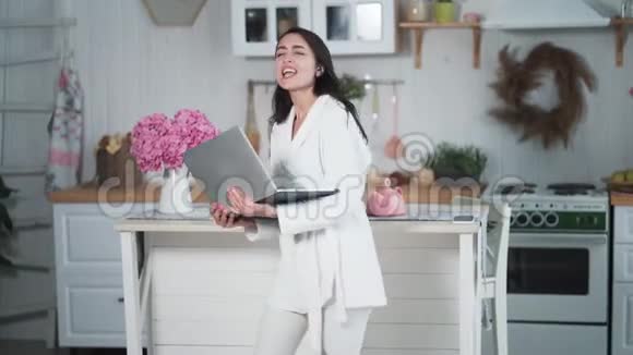 穿着浴袍的年轻女孩在家里的厨房里用笔记本电脑工作唱歌跳舞视频的预览图
