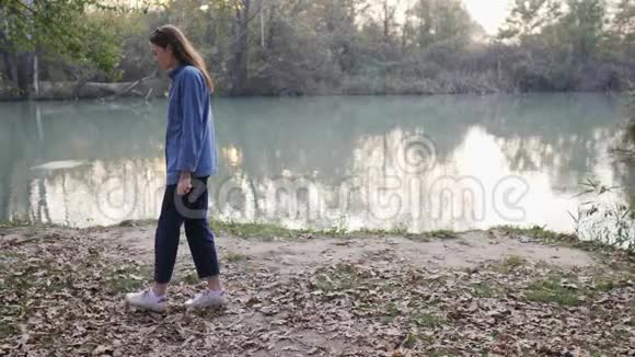 一位美丽的年轻女子在森林背景下在池塘附近的落叶上随意散步秋天视频的预览图