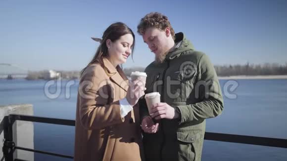 在河岸边爱喝咖啡的年轻夫妇的肖像快乐的白人男女在阳光明媚的户外约会视频的预览图