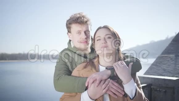 年轻幸福夫妇看着相机在户外微笑的肖像可爱的白人男女在河上摆姿势视频的预览图