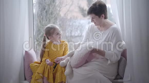 快乐的白人祖母和孙女在室内窗台上聊天的肖像微笑的成年女人和小女孩视频的预览图
