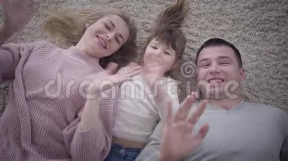 高瞻远瞩的特写镜头微笑的高加索家庭躺在柔软的地毯上对着镜头挥手快乐父亲的画像视频的预览图