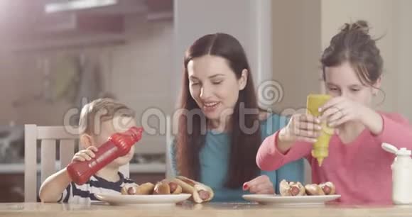 妈妈正在教她的孩子做热狗因为他们正在学习用红镜头拍摄厨房餐桌上的热狗视频的预览图