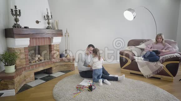 笑着一个金发白种人女人坐在扶手椅上拿着平板电脑看着她的丈夫在玩四弦琴和女儿视频的预览图