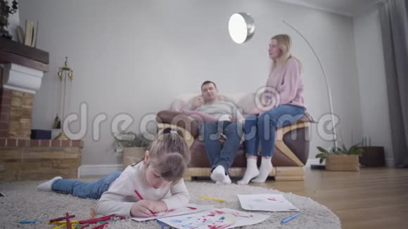 一对开朗的白种人年轻夫妇坐在扶手椅上聊天正当他们可爱的小女儿在前台画画视频的预览图