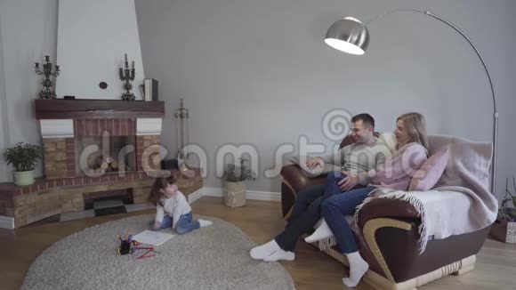 快乐的白种人已婚夫妇坐在扶手椅上聊天他们可爱的小女儿在地板上画画视频的预览图