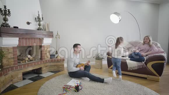 白人父母欣赏他们欢快可爱的女儿在室内跳舞和旋转的肖像微笑的母亲坐着视频的预览图
