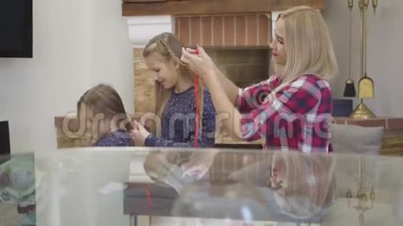 侧面看着金发白种人的母亲和两个女儿互相编织头发在玻璃桌上思考的人视频的预览图