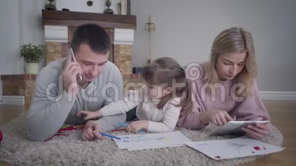 可爱的小白种人女孩和父母一起躺在地板上画画的肖像年轻的父亲在打电话视频的预览图