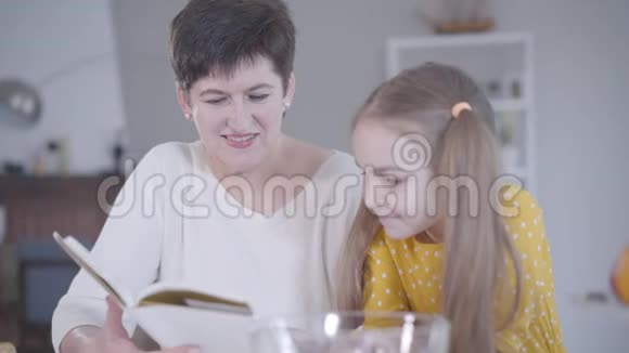 微笑的白种人祖母在室内给孙女读煎饼食谱的肖像快乐的成年女人视频的预览图