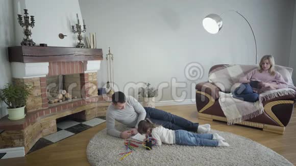 年轻的白人父亲和女儿躺在地板上的软地毯上母亲坐在扶手椅上画画视频的预览图