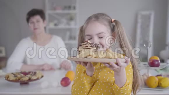 快乐的小白种人女孩闻着烤薄饼把盘子和甜点送给微笑的祖母的肖像视频的预览图