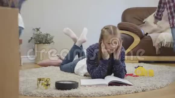 可爱的高加索女学生躺在柔软的地毯上当她的弟弟妹妹四处奔跑时阅读的肖像小可爱视频的预览图