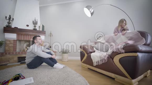 侧面观察年轻的白人男子坐在壁炉前看着他的妻子和女儿玩捉迷藏视频的预览图