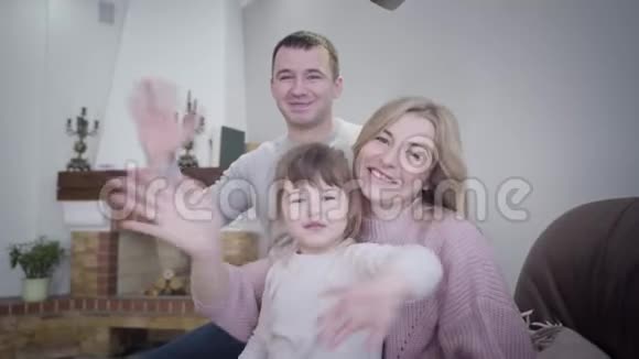 快乐的白人家庭在镜头前挥手的肖像微笑的男人女人和坐在扶手椅上的小女孩视频的预览图