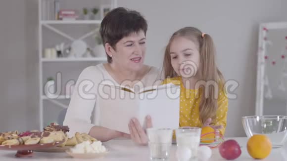 在厨房里读食谱的漂亮女孩和成年女人的肖像快乐的高加索祖母和孙女视频的预览图