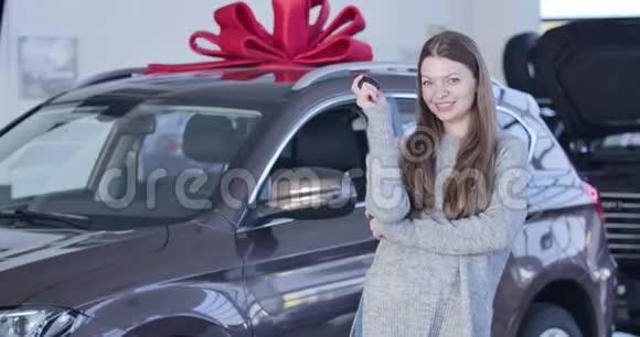 年轻的白人女孩吹嘘汽车钥匙旁边的新车与大红色弓在经销商快乐成功视频的预览图