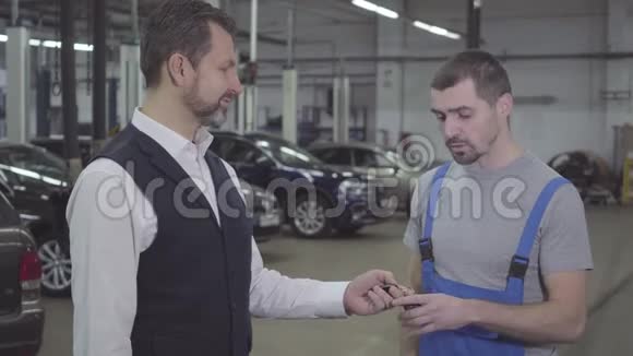 侧面观看英俊的成年白种人男子给汽车钥匙的汽车机械师和握手有自信的家伙视频的预览图