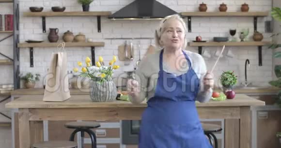 快乐的高加索女人在现代厨房里和烹饪设施跳舞欢快的中年退休人员画像视频的预览图