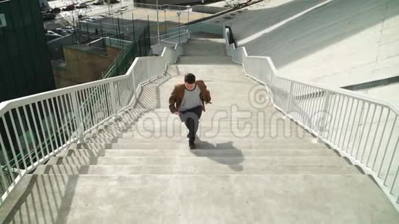 快乐的年轻人上楼梯时积极跳舞他穿一件棕色的长外套在城市公园拍摄的场景慢动作视频的预览图