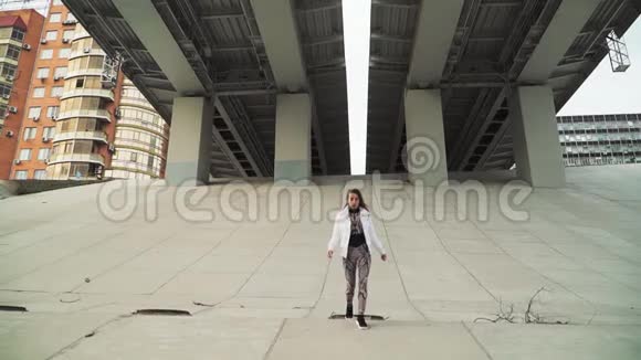一个穿着白色夹克的年轻漂亮的白人女孩在一座大城市桥下的水泥斜坡上慢动作视频的预览图