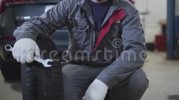 不可辨认的白人男子穿着工作服坐着汽车车轮和扳手并显示拇指男汽车修理工视频的预览图