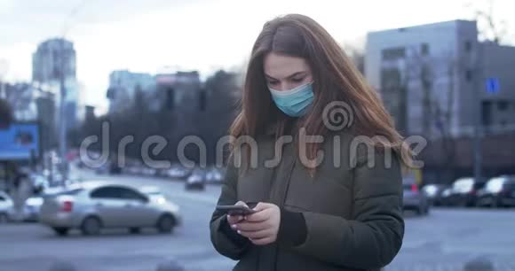 忧心忡忡的女人戴着防护面具站在城市街道上并使用智能手机的肖像忙碌的女孩在看新闻视频的预览图