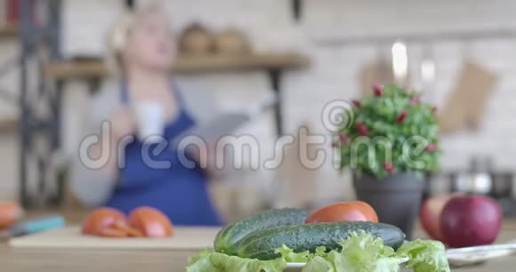 厨房的餐桌上摆着新鲜蔬菜的特写镜头模糊的女人一边喝着咖啡一边在厨房看菜谱视频的预览图