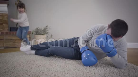 快乐的小白种人女孩跑到年轻人面前戴着拳击手套躺在柔软的地毯上女儿拳击视频的预览图