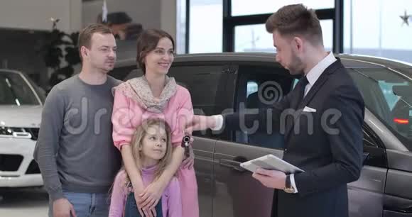 可爱的白种人小女孩拿着车钥匙从经销商父亲抚养她四个人看着摄像机微笑着视频的预览图