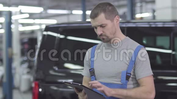 年轻英俊的白种人穿着工作服站在黑色汽车的背景下拿着平板电脑严肃的男汽车修理工视频的预览图