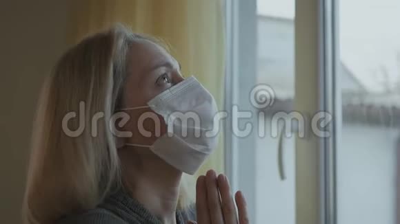 他脸上戴着医用面具的特写女士用双手交叉在窗户前祈祷视频的预览图