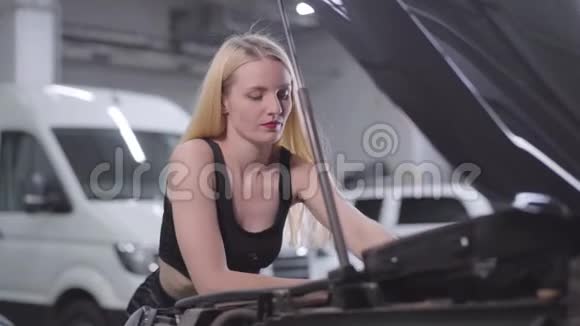 镜头在美丽的金发白种人女人周围移动在打开的汽车罩上拧紧螺丝性感女汽车修理工视频的预览图