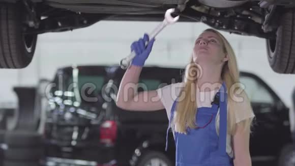 年轻白种人女人站在车下拧紧螺丝的肖像蓝色工作服的女汽修工视频的预览图