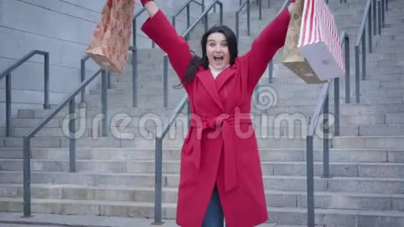 穿着红色外套带着购物袋在户外跳舞的快乐白人女孩的肖像令人兴奋的女购物狂满意视频的预览图