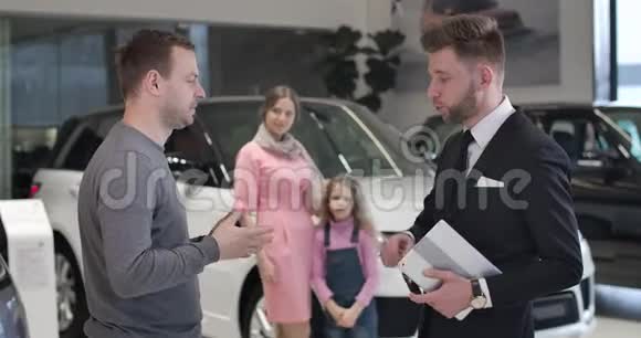年轻的白种人商人说着车钥匙和颤抖的商人他的手作为他幸福的妻子和女儿拥抱在视频的预览图