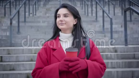 欢快的年轻女子喝着咖啡举着杯子对着镜头的肖像穿红色外套的漂亮女孩视频的预览图