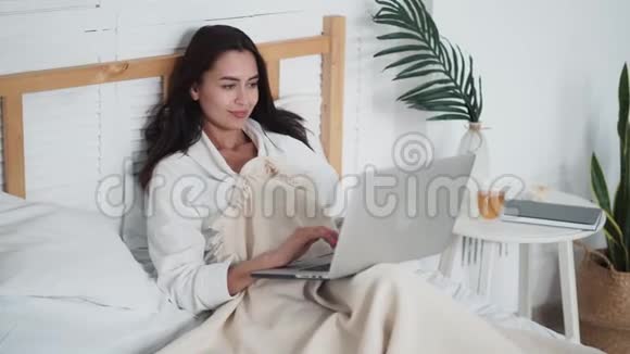 美丽的年轻女子躺在床上用笔记本电脑工作或在线聊天视频的预览图