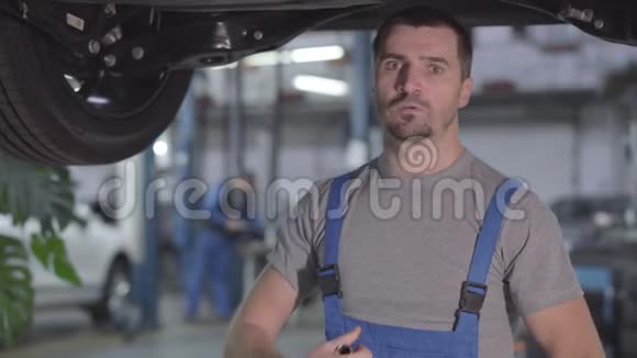 年轻严肃的白人男子穿着蓝色工作服站在修车厂的车下看镜头的肖像视频的预览图