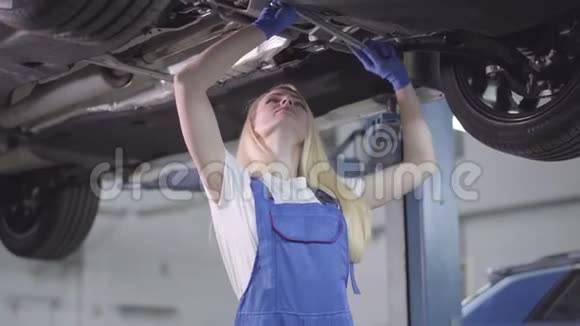 一个年轻自信的白种人女人穿着工作长袍站在车下拧紧螺丝美丽的女人视频的预览图