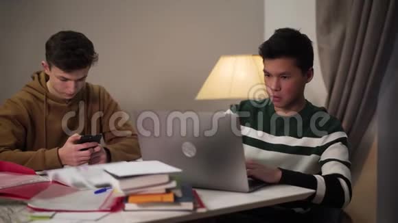 集中的十几岁的亚洲男孩展示笔记本电脑屏幕给他的高加索队友使用智能手机两个帅哥视频的预览图