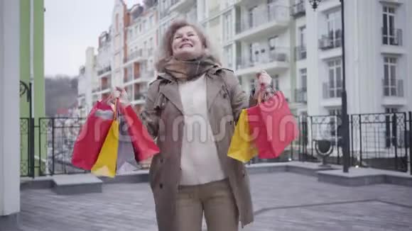 满意的高级女性购物狂用购物袋在城市街道上旋转和跳舞的肖像成熟的白种人视频的预览图