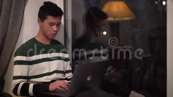 年轻的亚洲男孩在笔记本电脑上打字和晚上看窗外的肖像帅哥大学生视频的预览图