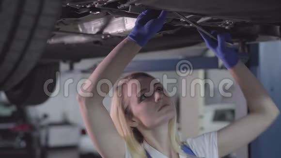 年轻的白种人女人站在车下拧紧螺丝在修理厂工作的金发女汽修工视频的预览图