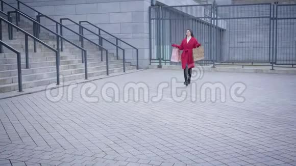 年轻快乐的白种人女人穿着红色的外套和高黑色的靴子沿着城市广场走着离开满意的女孩视频的预览图